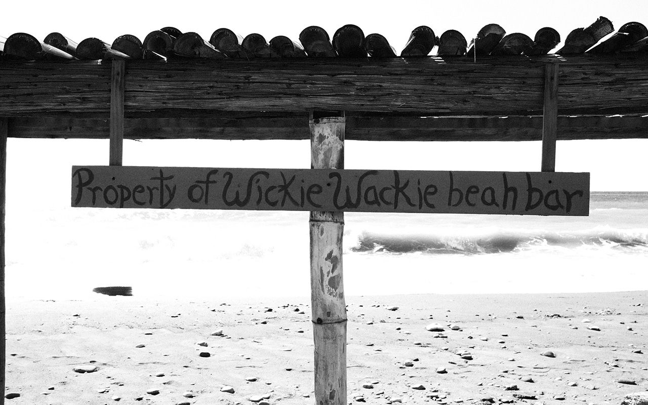 Photo of a thin, horizontal, hand made wooden sign on the sandy shore of a beach with the words "Property of Wickie Wackie beat bar" hand-painted in fancy writing. The sign is nailed onto a bamboo post and supported on top by a wooden beamed "roof" lined with some more bamboo posts. Waves rolling onto the shore are visible in the background.