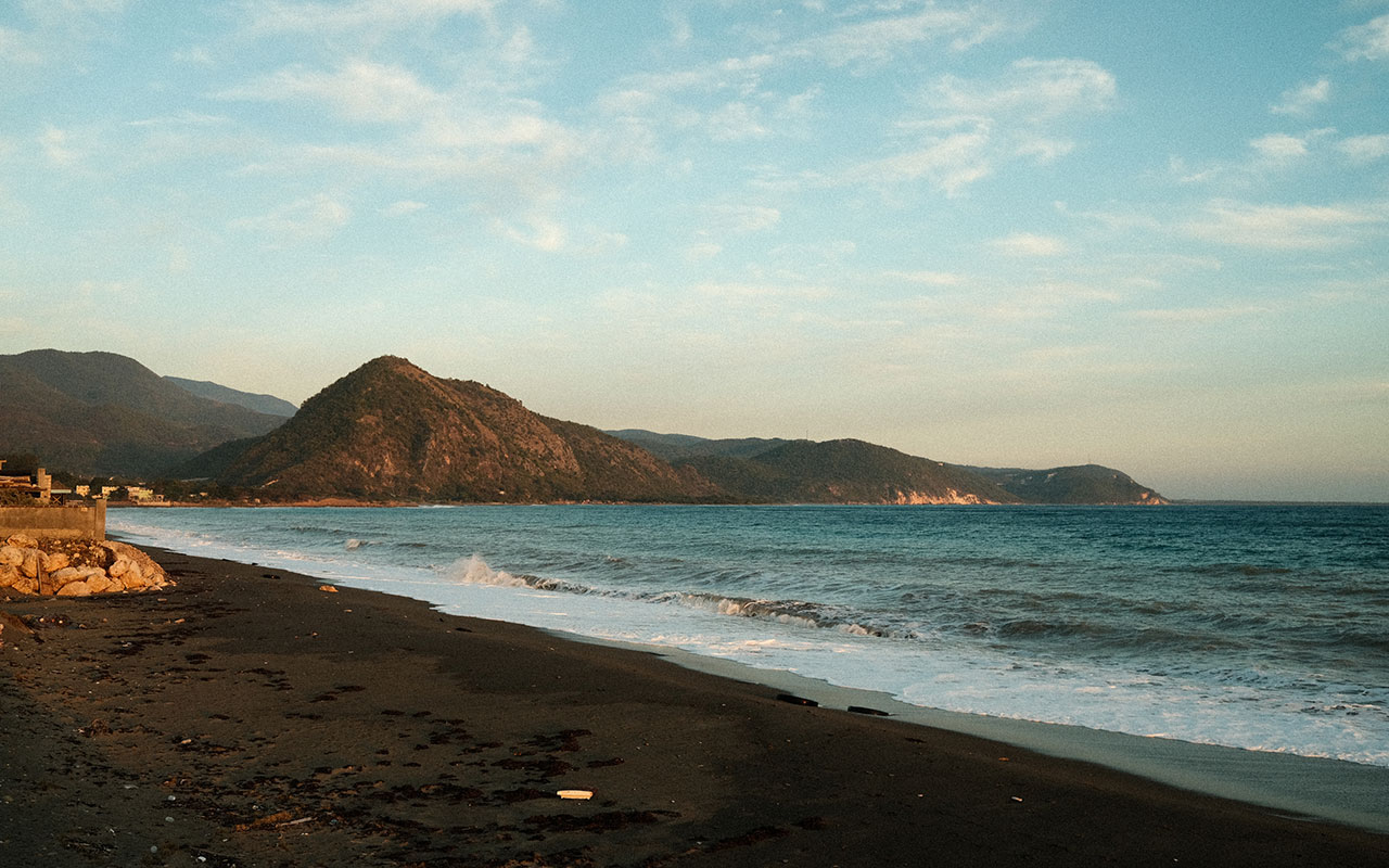 A photo of a dark sand beach in Bull Bay, Jamaica taken from the shore just before sunset. The water is a deep turquoise blue and dark green tree-covered mountains are in the distance.