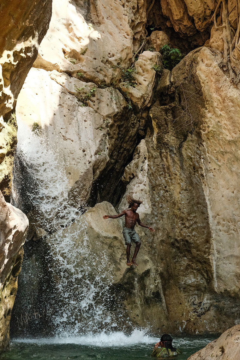 Action shot of a man jumping from a small ledge carved in the surrounding light brown, sand-colored rocks. The man is jumping into the small alcove of light green water below that appears tp be filled by a small waterfall. Another person is wading in the water below.