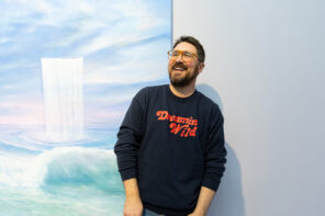 Jeremy Shockley in a black shirt with red lettering posing next to his painting hanging on the wall at his art show. The painting is oil on canvas and depicts a light green ocean beneath a cloudy purple and blue sky. There is a sheet-line waterfall pouring vertically out of the sky into the ocean below.