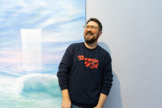 Jeremy Shockley in a black shirt with red lettering posing next to his painting hanging on the wall at his art show. The painting is oil on canvas and depicts a light green ocean beneath a cloudy purple and blue sky. There is a sheet-line waterfall pouring vertically out of the sky into the ocean below.