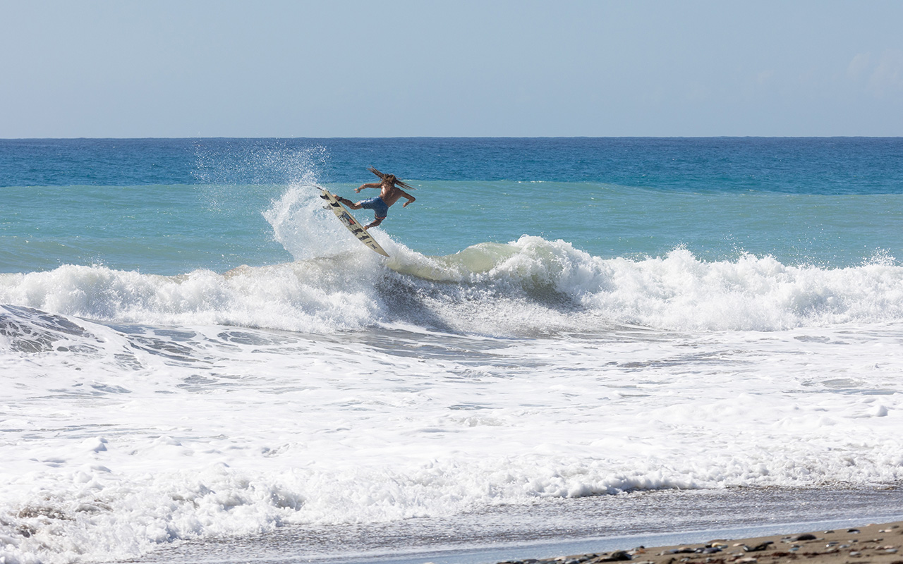 Photo of a man on an off-white surfboard jumping a wave in the white water of the crystal blue ocean close to the sandy shore.