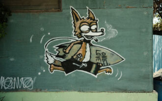 Close-up photo of a cartoon orange fox with a cigarette dangling from its mouth wearing black boardshorts and running with a surf board under its arm that has the words "ONE LOVE" scrawled onto it. The wooden wall behind the street art is painted jade green and the artist's white signature is spray-painted on the bottom left corner of the wall.