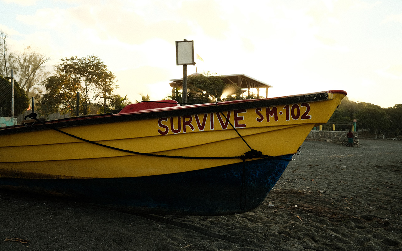 Photo of the mast of a black and bright yellow fishing boat with the boats name, "SURVIVE SM.102" painted in all capital dark red letters. The boat is docked on the sand. 