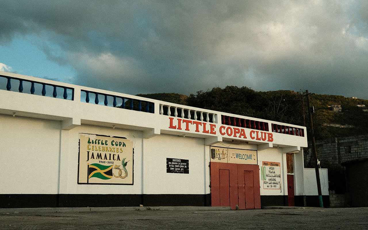 Photo of a clean white dement restaurant building with a sign that breads "LITTLE COPA CLUB" in red lettering and terracotta red double doors at the entrance Small signs, including one with the green, yellow and black Jamaican flag on it, are posted on the front of the restaurant.