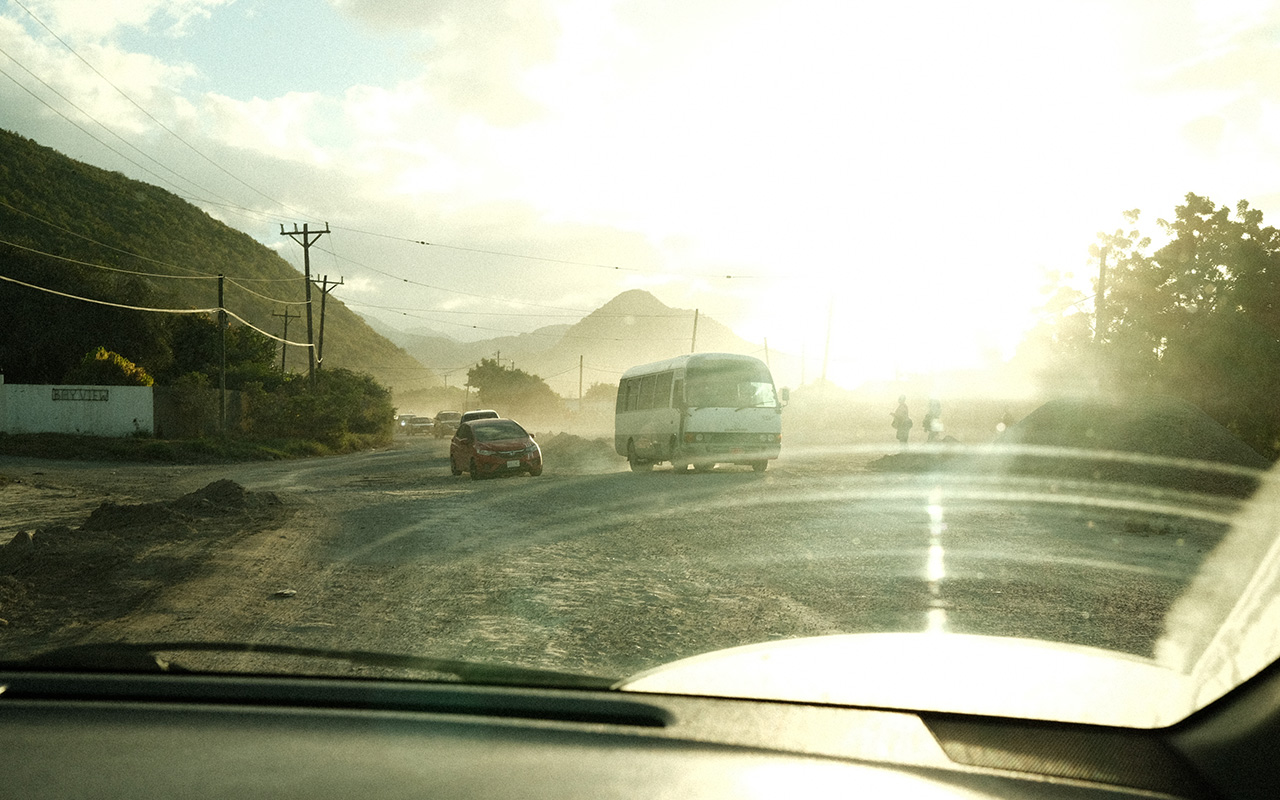 Photo taken at from the front windshield of a car of a dirt road and incoming traffic. Light yellow rays from the sun are shining brightly into the window, illuminating the smears created by the windshield wipers. The sun is setting against the mountain in the distance.
