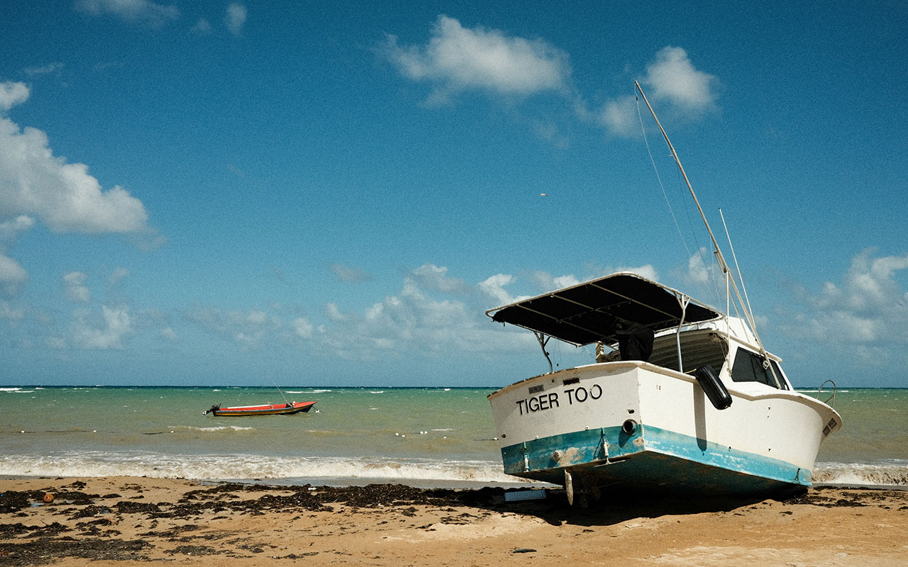 Photo of a white fishing boat with a light blue underbelly and a small awning parked on the shore of a light brown sandy beach. A small orange rescue boat is floating in the greenish blue ocean water close to the shore. The sky is bright blue with a few puffy white clouds.