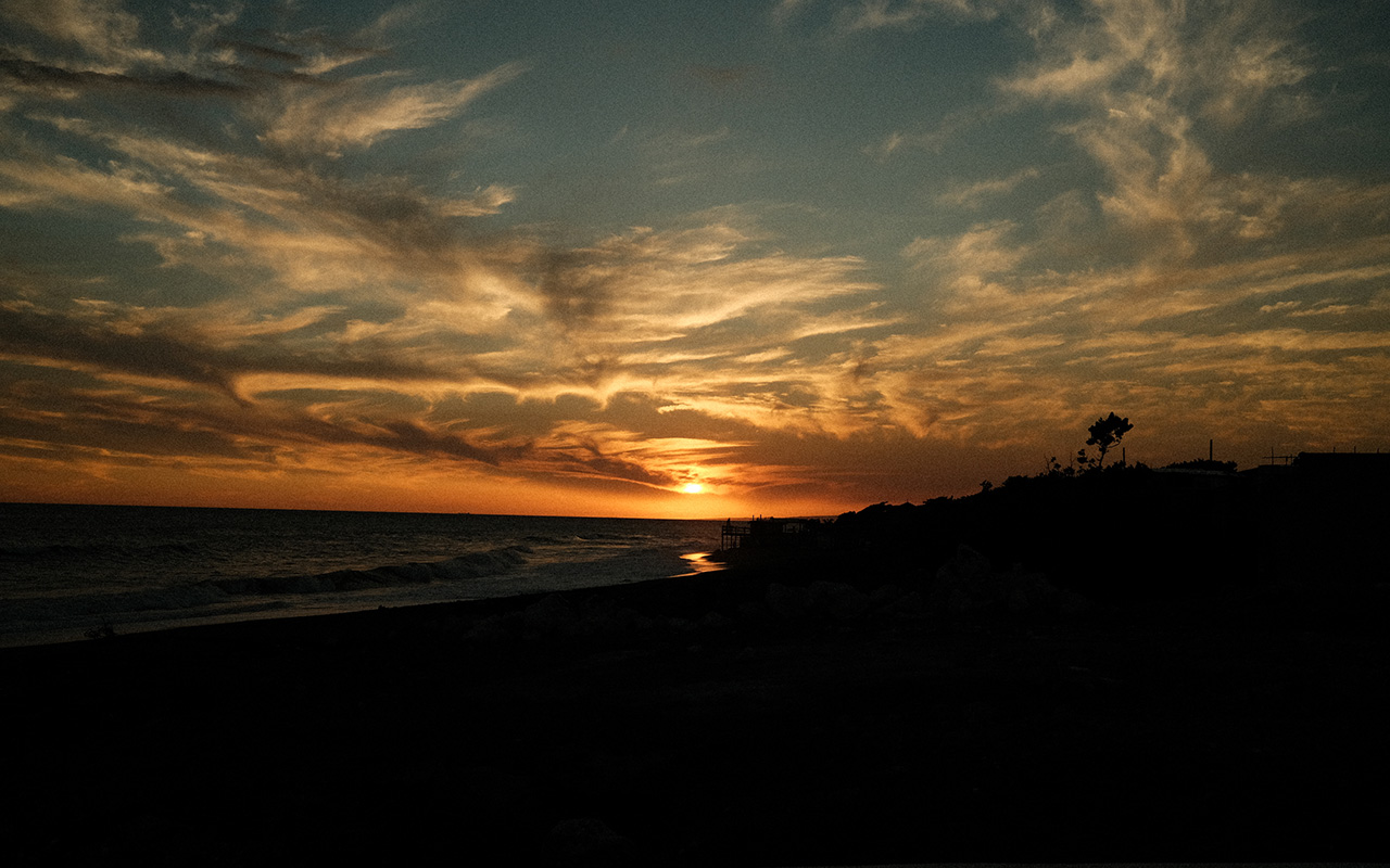 Photo taken on the sandy shore of a beach with the ocean to the left of the last light of day as the burnt orange sun sets in the distance and illuminates the horizon of the fading blue, partly cloudy sky.