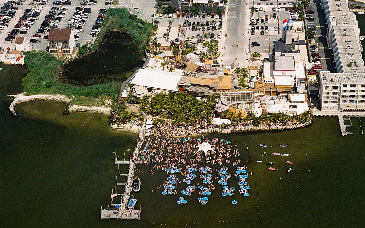 An aerial photo of the Seacrets, Jamaica resort in Ocean City, Mary.and. There are clusters of bright blue inner-tubes, jet skis and people to the right of the long wooden dock in the murky green water. Pal m trees line the shore of the resort and back up tonight gray buildings and parking lots.