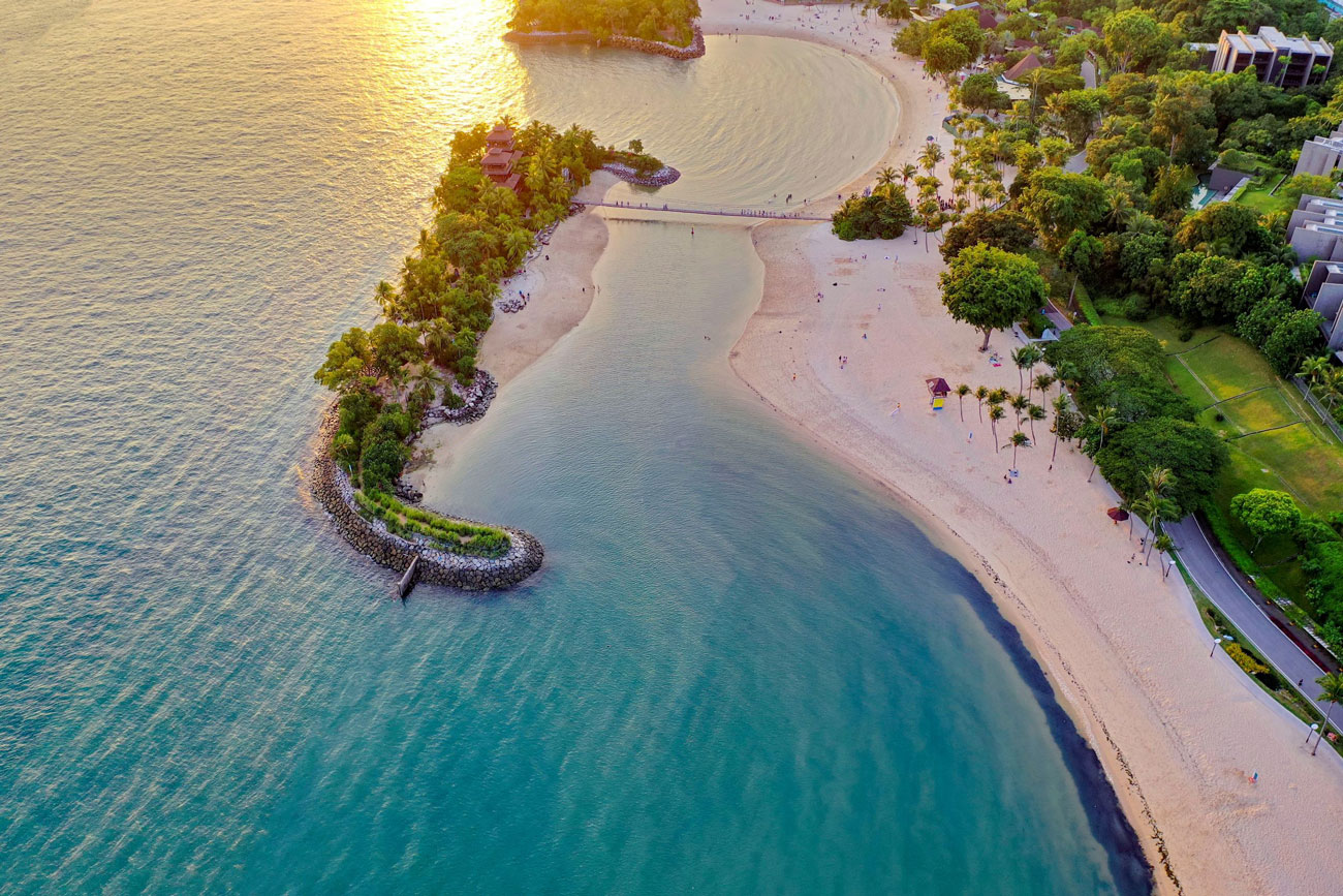 A small island in the middle of crystal blue waters. The island is in a backwards J shape with rocks on the shoreline and is covered with palm trees. The island back up to the sandy beach of the mainland and is connected via a thin bridge.