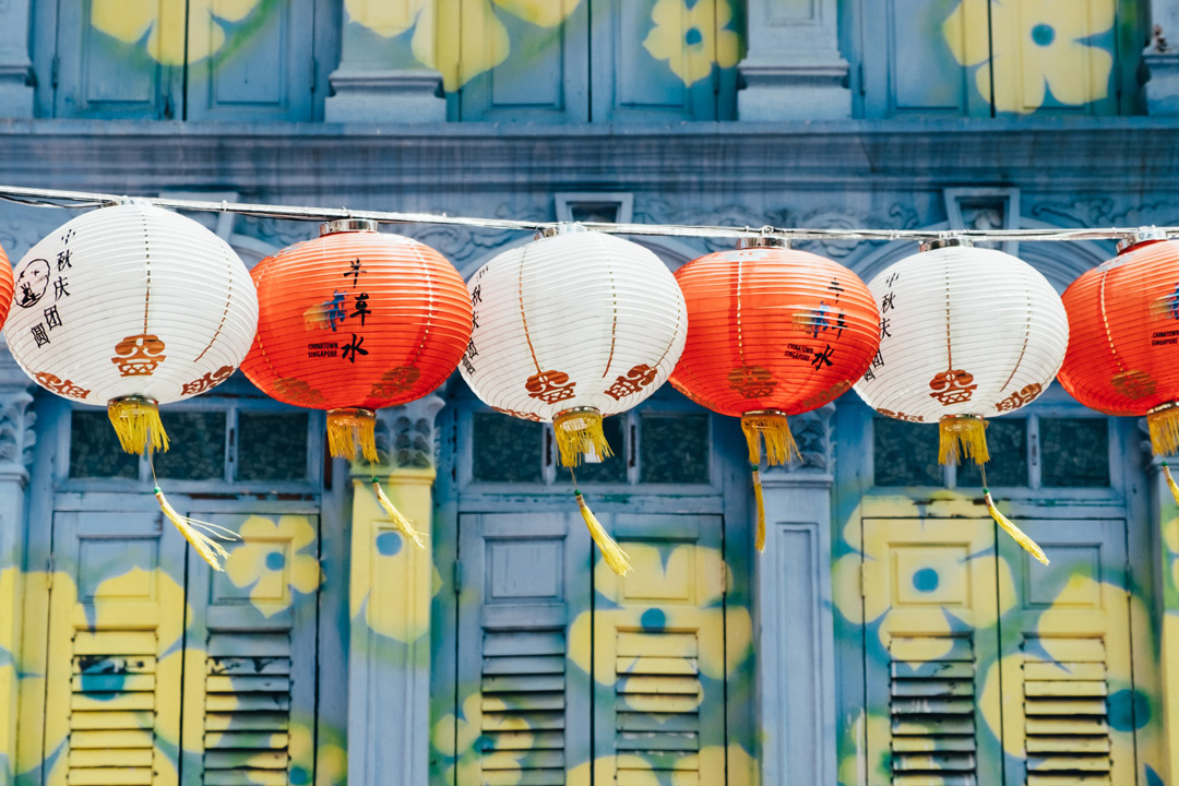 Red and white Chinese lanterns hanging on a line reaching across the photo. The wall behind the lanterns is a light blue color with yellow flowers painted on it.