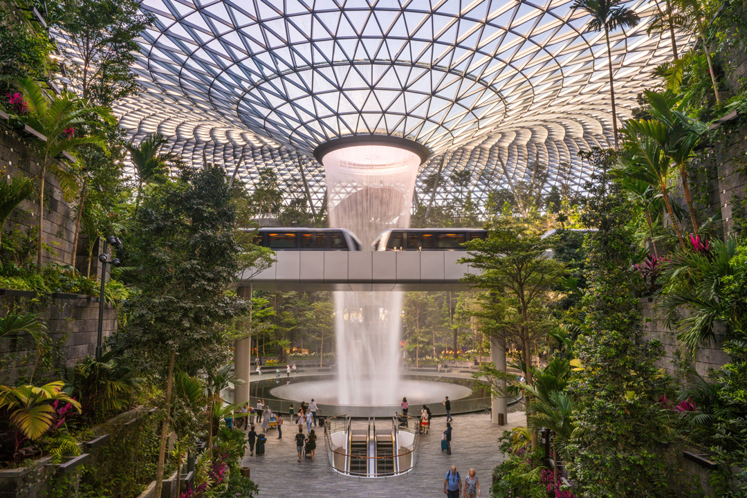 Changi Airport indoor waterfall. The middle of the large room has a glass ceiling with a hole in the middle that has water falling through it into a pool below. The sides of the building are all covered in trees and plants making the inside look like a large garden. There is an airport tram speeding across a rail line in the air. People are walking around the ground level with their suitcases and luggage.