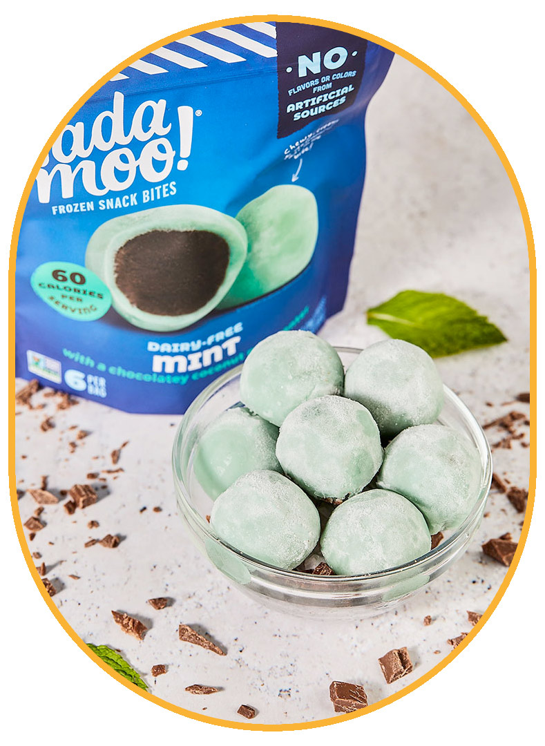 Glass bowl filled with light green Mint Nadamoo! Snack Bites. Cut up chocolate and mint leaves are scattered decoratively around the bowl and a royal blue package of NadaMoo! Mint Snack Bites is visible in the background. 