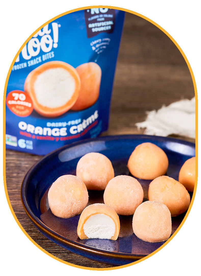 NadaMoo! Orange Crème Snack Bites (light orange exterior, white interior) sitting neatly on a dark blue ceramic plate. A royal blue package of the Snack Bites is visible in the background on the dark brown wooden table. 