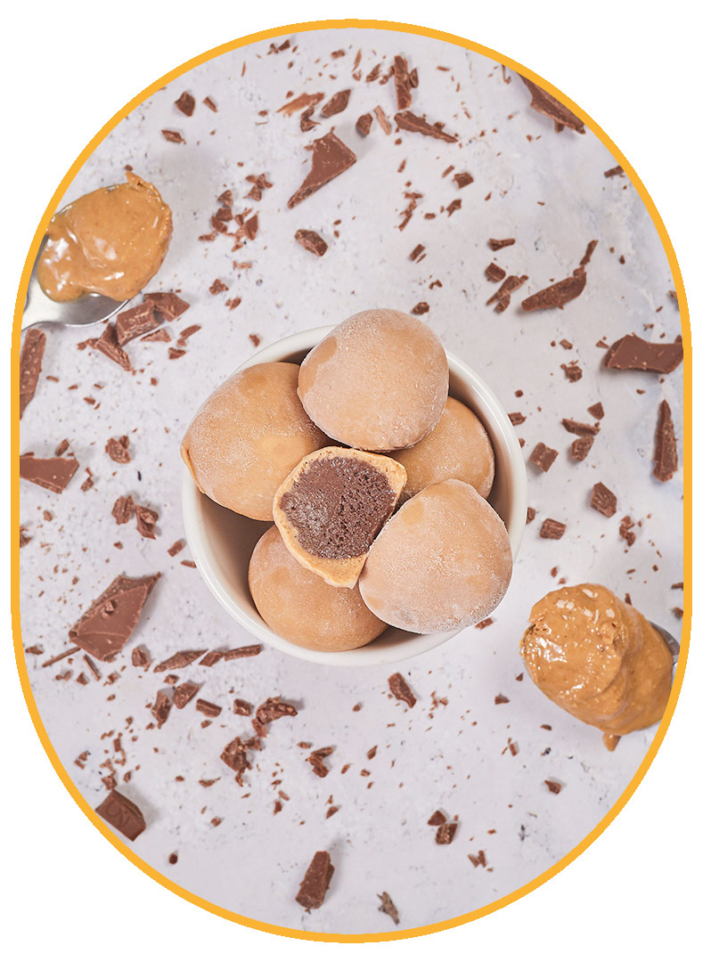 Aerial view of a bowl of light brown Peanut Butter NadaMoo! Snack Bites sitting on a white marble table. One Snack Bite is cut in half to expose the dark brown interior. Silver spoons full of peanut butter are hovering on either side of the bowl and bits of chopped chocolate are scattered on the table. 