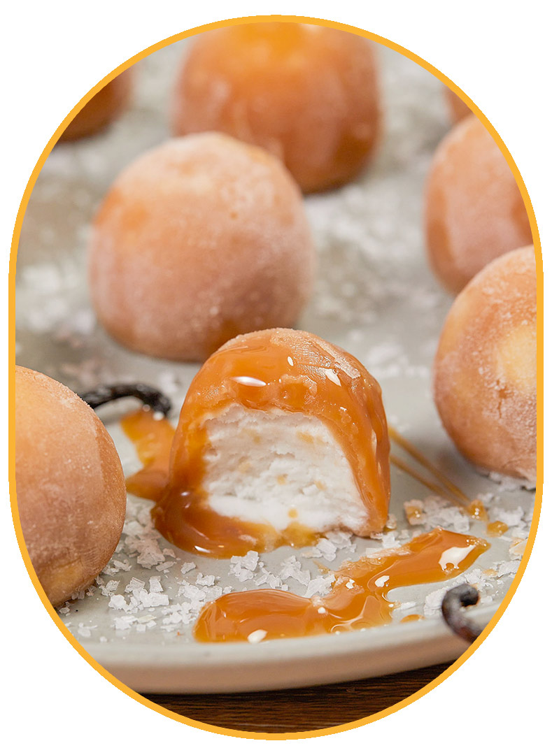 Closeup of a Salted Caramel NadaMoo! Snack Bite with a bite taken out of it. The exterior is light brown and the interior is while. The Snack bite is sitting on a plate of other snack bites decorated with lots of sea salt. 