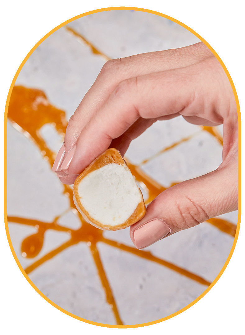 Aerial photo of a hand holding a light brown Salted Caramel NadaMoo! Snack Bite with a bite taken out of it  to expose the white interior. In the back ground, the white marble table is covered in light brown caramel drizzle. 