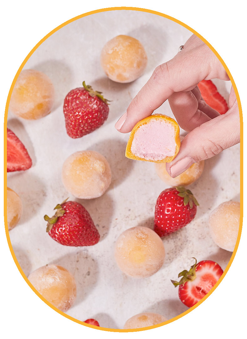 Close up of a hand holding a NadaMoo! Strawberry & Mango snack bite with a bite taken out of it. The Snack Bite is orange on the outside and light pink on the inside. In the background on t eh white marble table, the snack bites are neatly placed in alternating rows of fresh whole and halved red strawberries. 