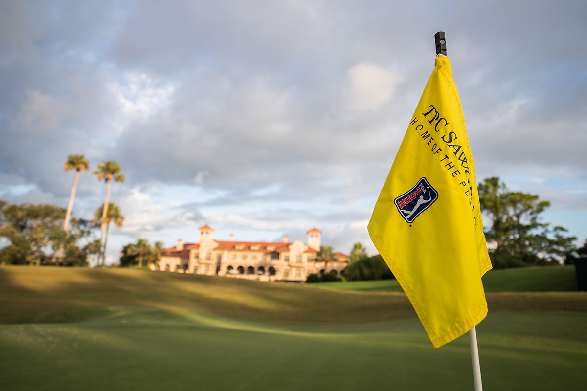 Close up photo of a bright yellow golf hole flag sticking out of the ground with the blue, white and red PGA TOUR logo on it and navy blue lettering that reads "TPC Sawgrass." and other words that cannot be mad out. A large orange-tiled roof and sandy colored mansion and the lush green golf course grounds of the Ponte Vedra Resort are blurred behind the flag.