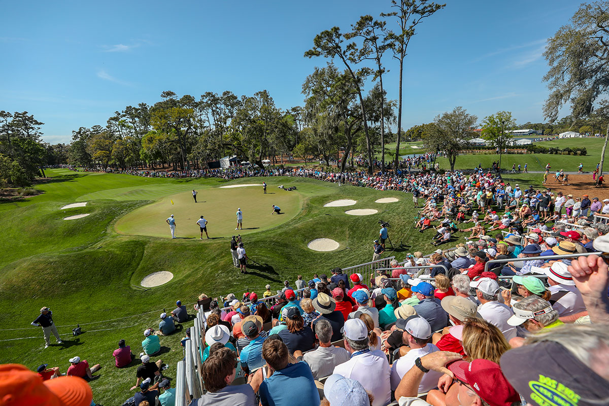 Photo taken from the stand filled with a large crowd of people of golfers standing on a lush green golf course around a hole during The Players Golf Tournament.