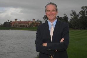 Photo of Jared Rice in a navy suit standing on the golf course next to one of the ponds at Ponte Vedra on a dark, cloudy day. Jared is smiling at the camera with his arms folded across his body.