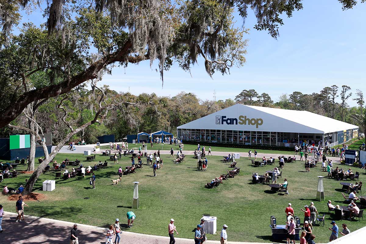Photo taken up high of a small, green park-like clearing at The Players Tournament. People are lounging in chairs, walking around and eating at tables. A large tent with a white awning with the PGA TOUR logo and gold and blue letters that read "FanShop" is behind the clearing .
