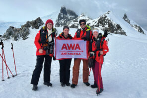 Four people standing in the snow smiling for a picture holding a rectangular banner that reads "AWA Antarctica" on it. The group are all wearing bright read snow jackets, long pants, and snow boots. They all have backpacks on their backs and have large cameras slung around their necks. There are red hiking poles sticking out of the snow on the left side and behind the group are snow covered mountains in Antarctica.