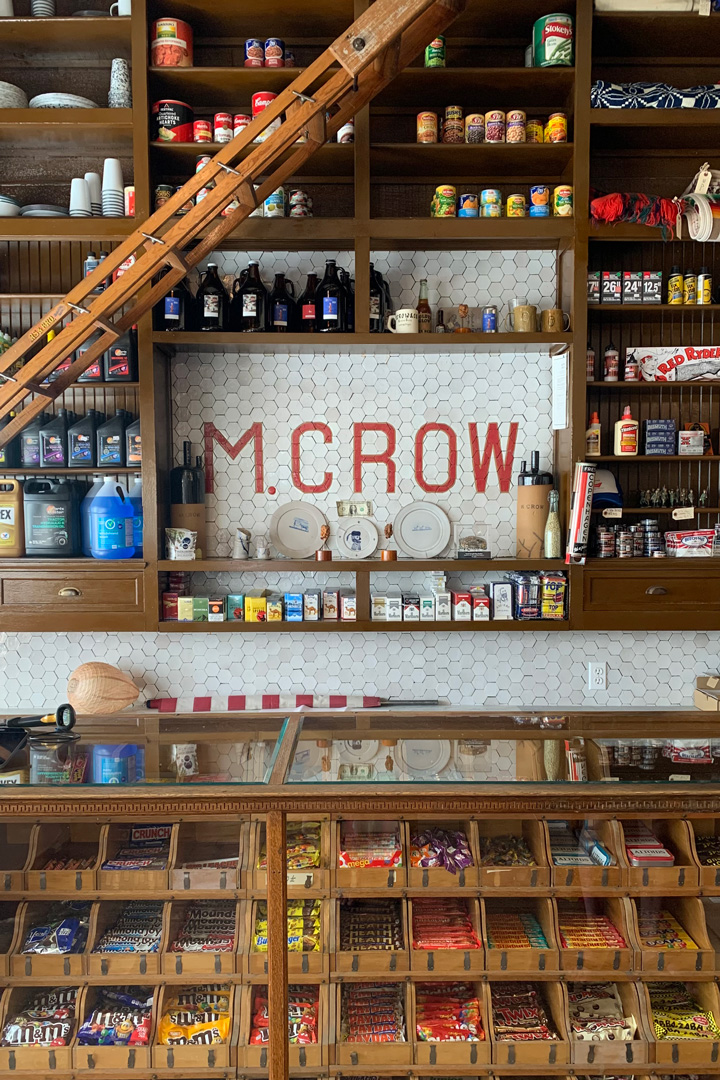 Photo of a wall in an old general store. The wall is lined with wooden shelves filled with various items; cigarettes, canned food, car oil, plates and cups, wood glue, and blankets. Right in the middle of the shelves is an opening to see the tiled wall behind that has the word "M. Crow" in red tile. There is wood cabinetry below the shelves that is filled with different types of candy; M&M's, Skittles, Butterfinger, Almond Joy, and Crunch bars amongst others. There is a small wooden ladder reaching diagonally across the picture in the top left corner.