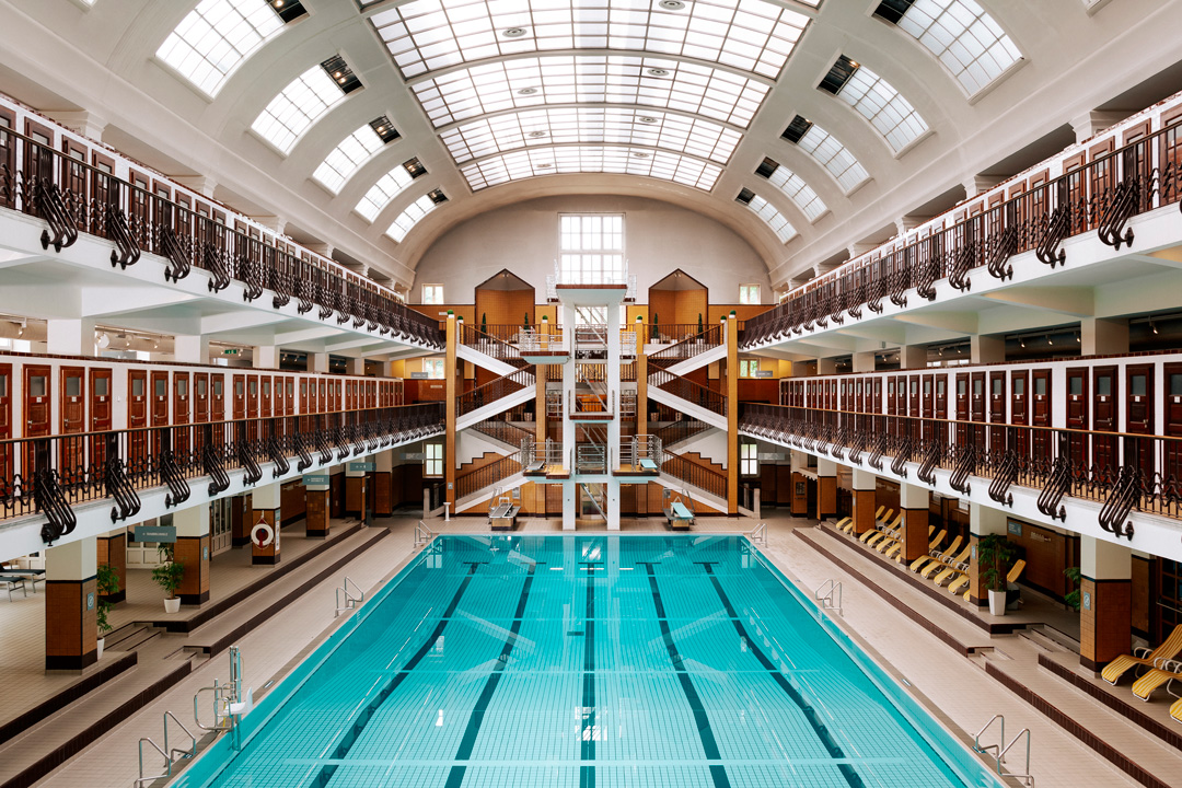 Very symmetrical photo inside of an indoor swimming pool. The building is very open and three stories tall with the pool in the middle. On the second and third floors on the right and left side of the pool have an iron railing and are lined with wooden doors all along the sides. The first floor by the pool has yellow reclining chairs on the right side and an open space on the left. There are two sets of staircases on the far wall that zig-zag up to the third floor. The pool is a bright cyan blue color and has lane lines at the bottom.