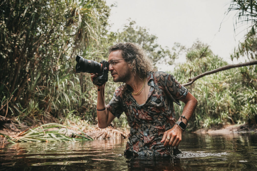 Photo of Donal Boyd standing waist deep in stagnant water in the middle of the murky green forests of Borneo. He is turned to the side and is looking through the viewfinder of the large black camera in his right hand.