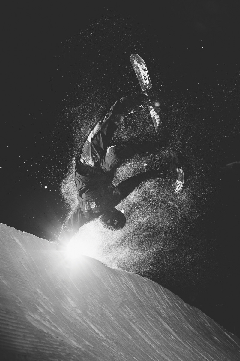 Black and white action shot of a person in snow gear holding the edge of a snow bank while flipping up side down in mid-air on a snowboard.