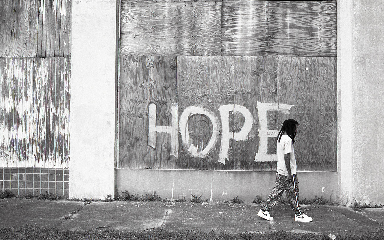 Black and white photo of a building with large windows boarded up with sheets of plywood. The word "HOPE" is painted in white on the plywood of the right window. On the right hand side of the photo just past the word "HOPE," a man with long black hair in casual clothing is walking the with head down and his hands in his pockets along the sidewalk overgrown with weeds in front of the building.