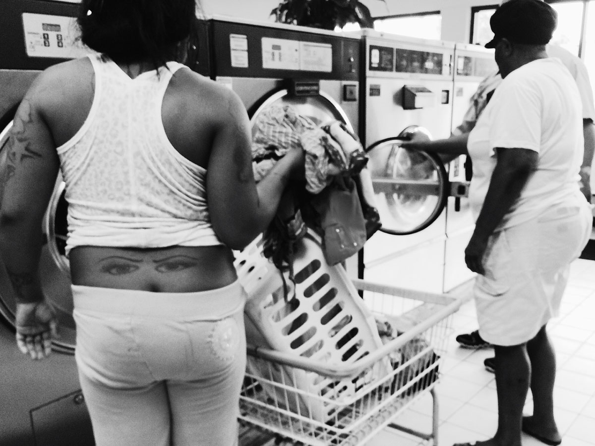 Photo of the back of two people loading their laundry from their rolling basket cart into a clothes washer at a laundry mat. The person on the left has a lower back tattoo of two eyes that look as if they are frowning at the camera.