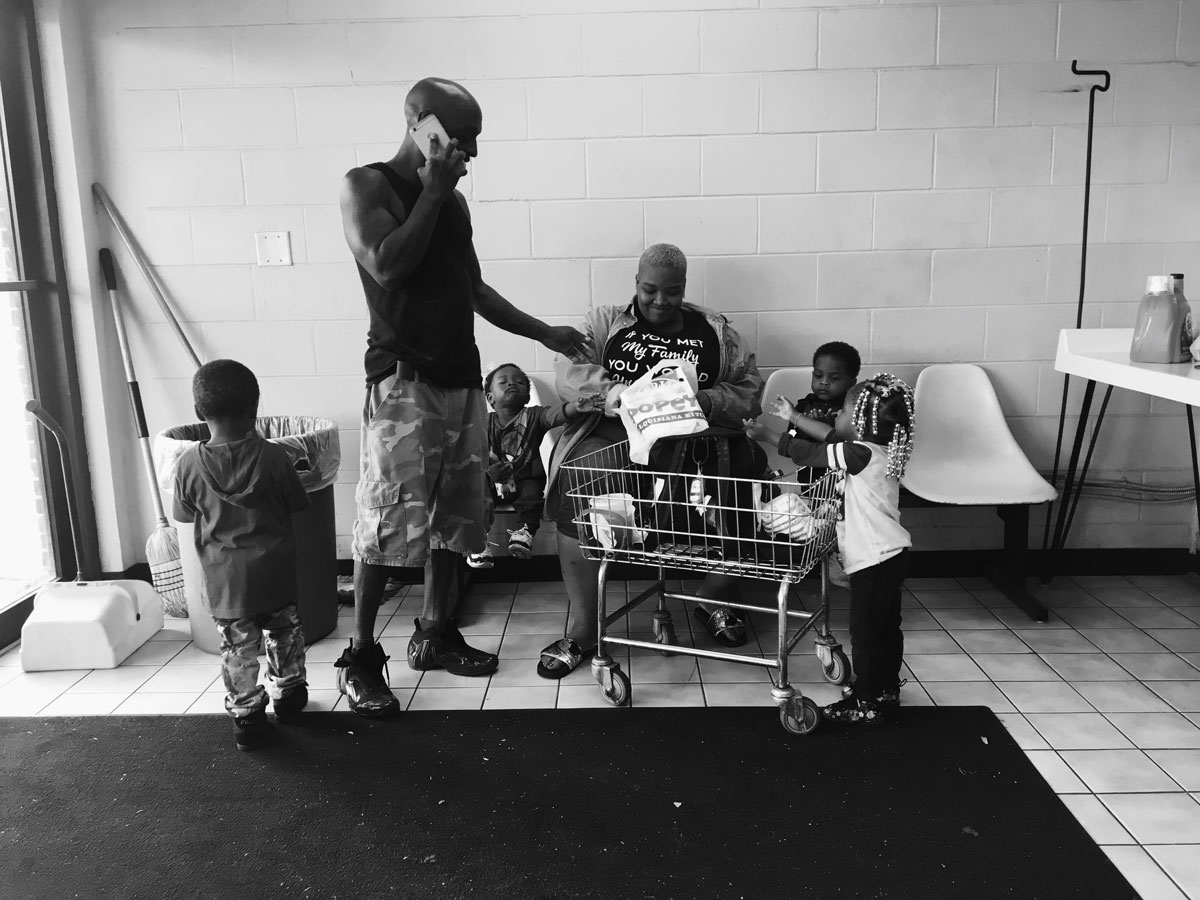 Photo of a family of four (a mother, a father and four young children) waiting at a laundry mat. The mother and two of the young boys are sitting in white chairs against the wall. The father is on the phone with one of the small boys standing to his left while the little girls rests her arms on the laundry basket cart in front of the father.