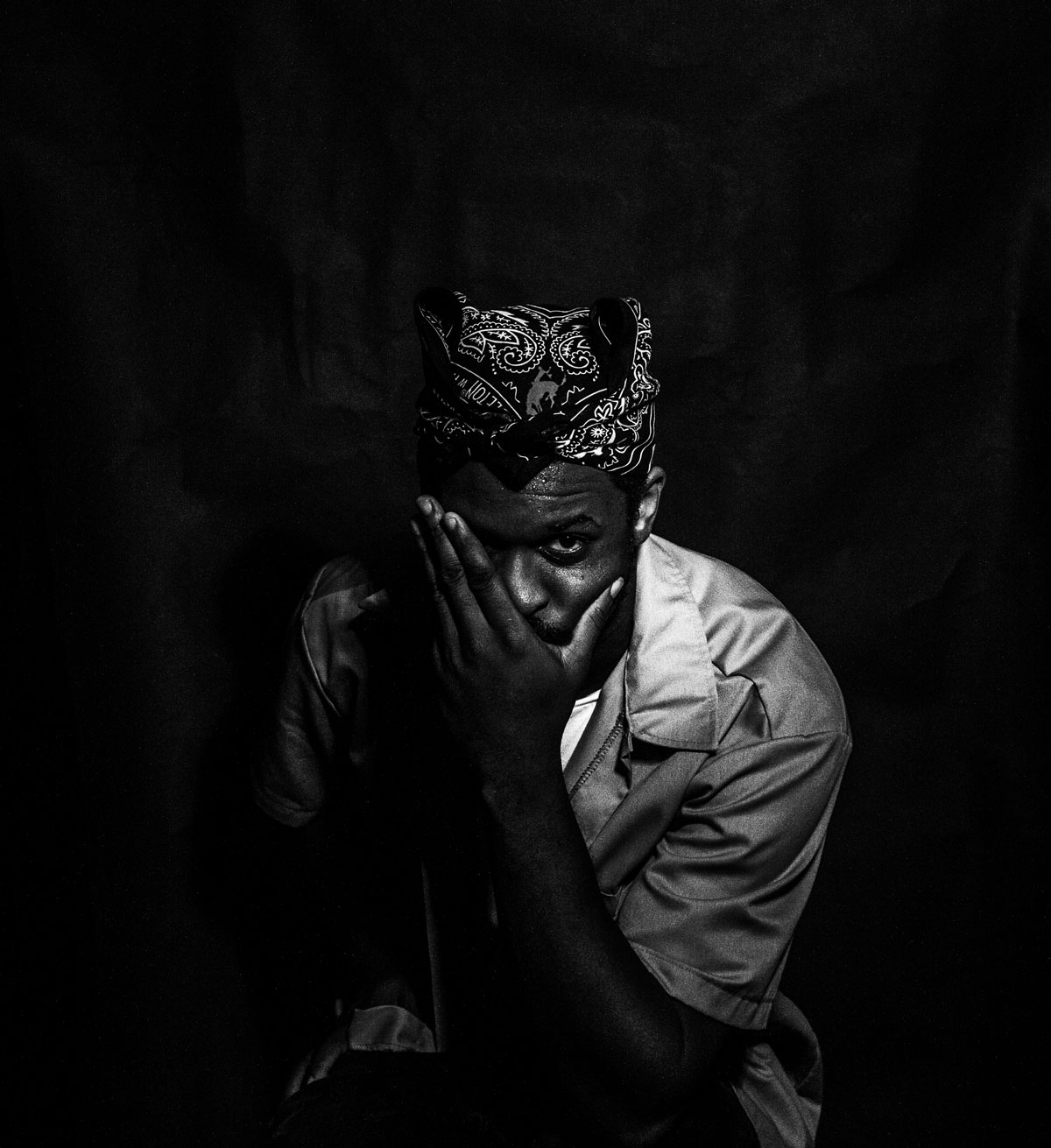 Portrait of a Black man wearing a bandana and a collared shirt. The man is covering the right half of his face with his left arm and looking slightly up at the camera with his uncovered eye. The man's face is covered in a slight sheen of sweat. The background of the photo is jet black. 