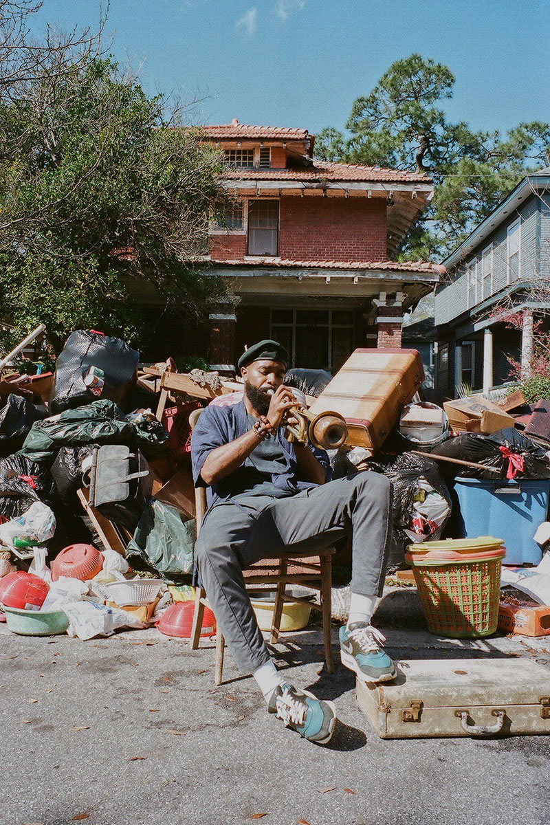 Photo of a man sitting back on a chair  in a driveway playing the trumpet. Behind the man is a large pile of discarded items and trash bags. Behind the pile of discarded items and trash is a large multi-floored red brick house.