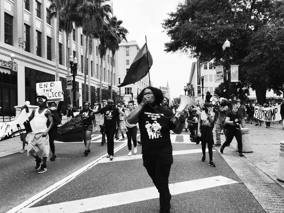 Black and white photo of protestors walking through a city street. The person closest to the camera is speaking through a megaphone. Other. protestors are holding handmade signs and flags as they march through the street. 