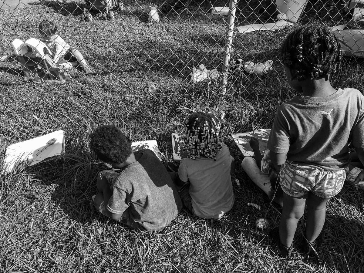 Black and white photo of the back of three children sitting in the grass drawing on drawing pads in front of them. A wire chainmail fence is separating them from another little boy sitting in the grass on the other side of it playing with a toy castle. 