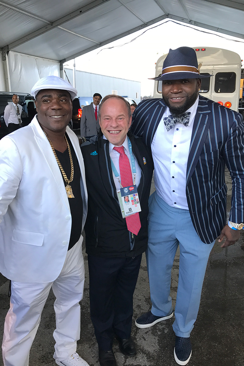 Photo taken at the Kentucky Derby of Tracy Morgan (left) wearing a white suit and matching white hat, Larry Collmus (middle) in a black Adidas jackets, slacks, a dress shirt and a pink checkered tie, and David Ortiz (right) in a pinstriped navy blue sports jacket, gray slacks, navy sneakers, a floral bowtie and a navy fedora with a light brown stripe at the rim. All of the men have their arms around each other in a row and are smiling for the camera.