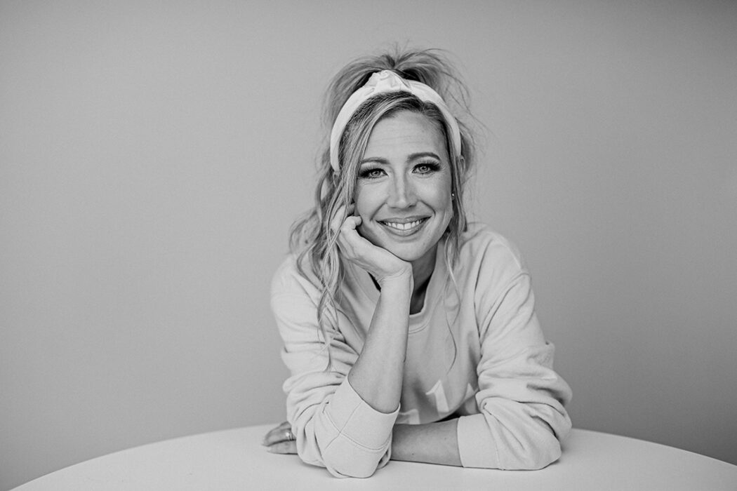 Black and white photo of Rebecca Reed leaning against a table and holding her chin in her hand while smiling. Her hair is tied up in a messy up-do and she has a white headband and a white long sleeve t-shirt on.
