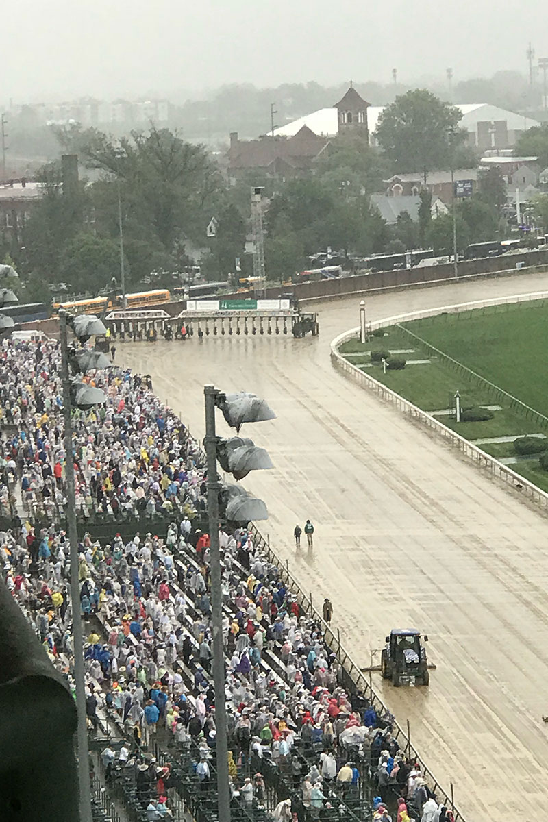 Photo taken from high in the stands of the empty tan racetrack and starting gates filled with horses lined up at the Kentucky derby. To the left, of the track, there is a packed crowd of people. It is a cloudy, rainy day and the track is glistening with water. 