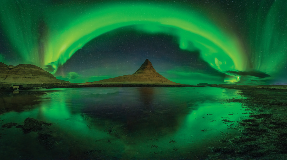 Panoramic photo taken on a calm lake of the bright green and yellow lights of the aurora borealis in the dark blue sky swirling of a large, pointed light brown rock formation in the distance. 