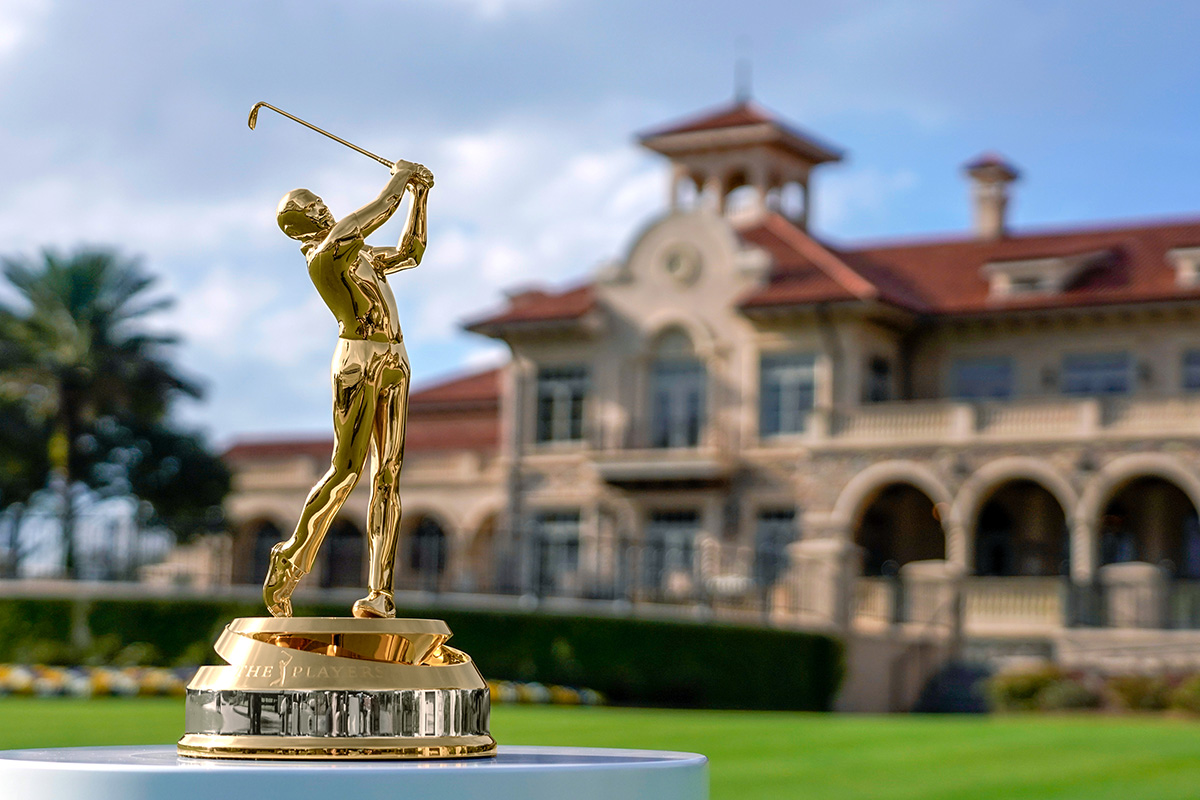 Close up photo of a gold trophy on a pedestal of a person swinging a gold club up behind their head. Behind the gold trophy is the large sandy brown foundation and clay-tiled roof of the mansion that sits on the bright green grounds of the Ponte Vedra resort where The Players Championship gold tournament.