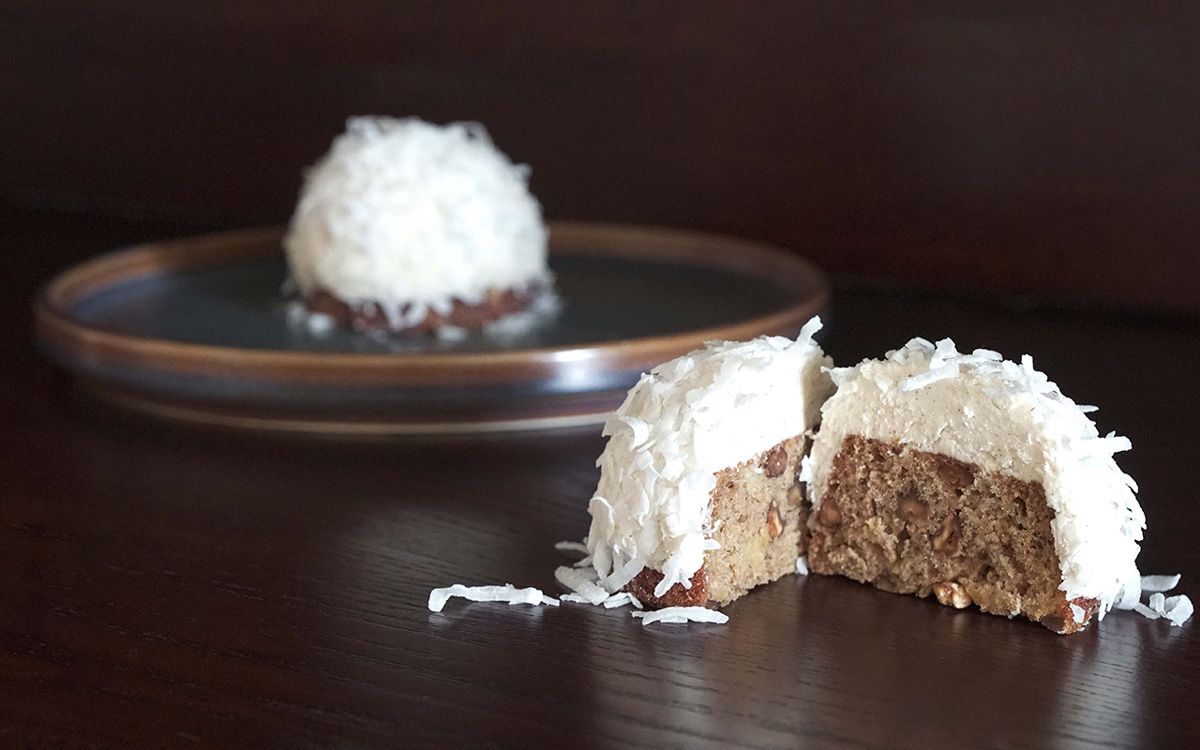 Photo of a two small cakes, one sitting blurred in the background on a plate and the other cut in half and focused unclose in the right foreground of the photo. The cakes are spherical with flat bases. The inside is light brown and the frosting is white with coconut flakes sprinkled on top.