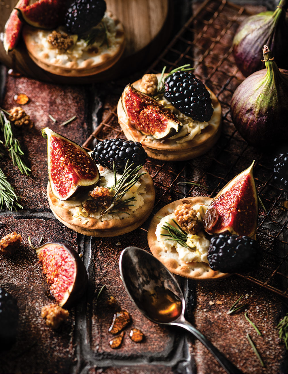 Close up photo of small circular matza-like cracker appetizers sitting on a dark brick surface. Each cracker is a light golden brown and is artfully decorated with hummus, small sprigs of green thyme, dark blackish purple blackberries, and quarter slices of bright pinkish red figs. 