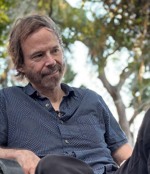 Photo of Author George Dawes Green lounging in a gray-blue shirt and dark gray pants on a chair in the middle of the woods surrounded by green trees.