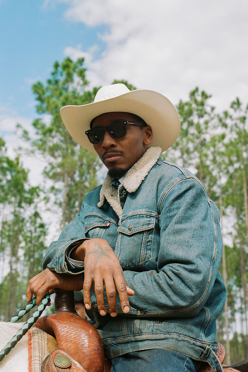 Close-up portrait of a Black man wearing a sherpa-lined jean jack, black opaque sunglasses and an off-white cowboy hat. The back of the white horse that the man is sitting on is just visible below the reigns that he is holding in one hand. The man appears to be looking at the camera. 