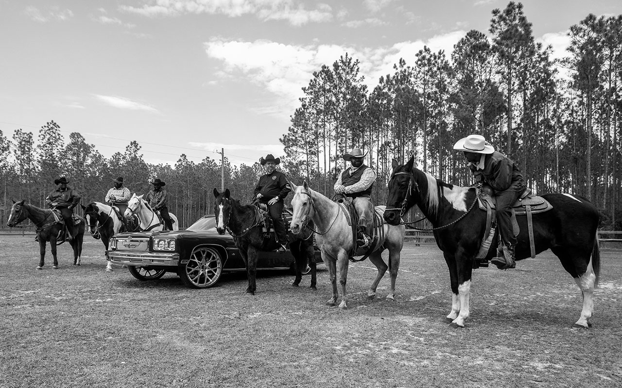 Black and white photo of 6 men in cowboy hats and western wear on horses talking to each other in a fenced in field. There is an old chevy Cadillac parked in between the men so that there are three men and horses on each side of the car. Tall, thin trees can be seen in the background.