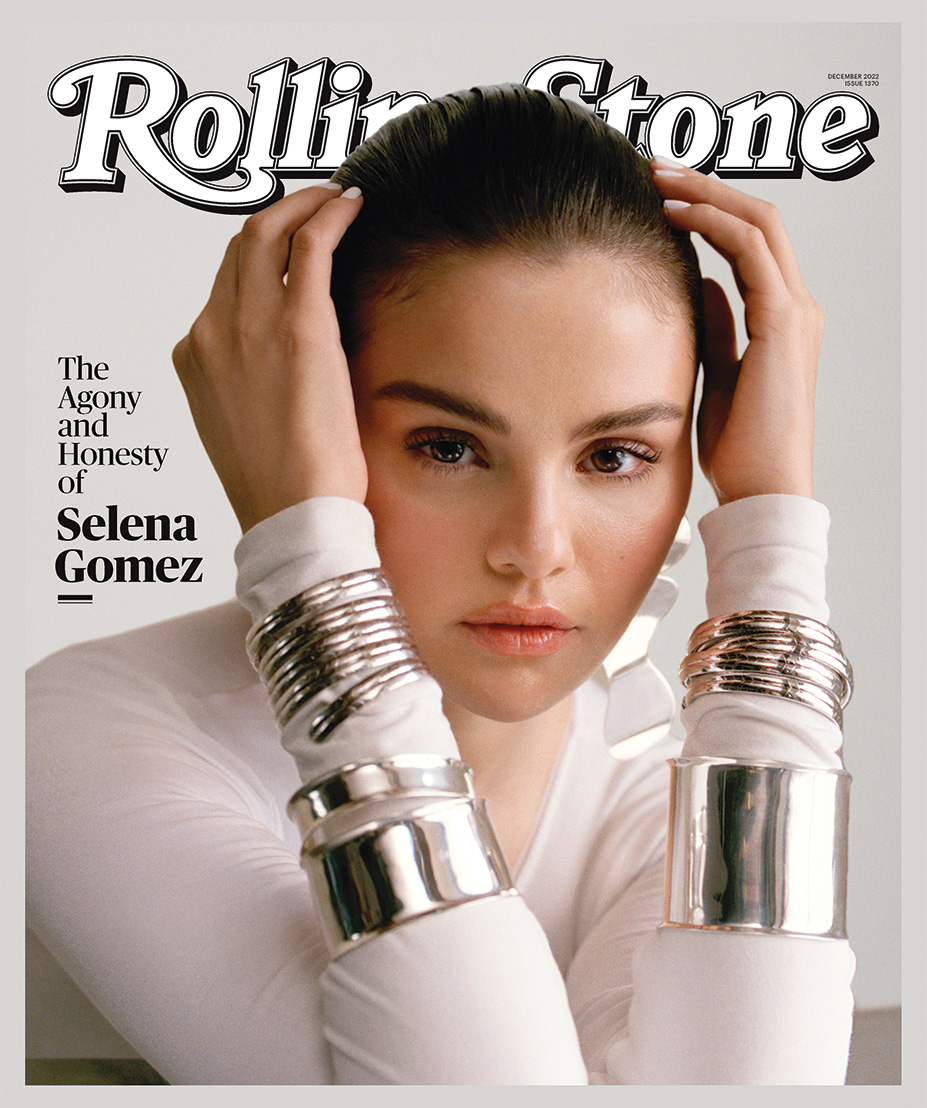 Rolling Stone magazine cover featuring a close up shot of Selena Gomez looking directly at the camera while leaning her elbows on a table with her hands covering the sides of her head. She is wearing a white long-sleeve shirt and large silver bangles around her wrists. The "Rolling Stones" title is white with a black outline.