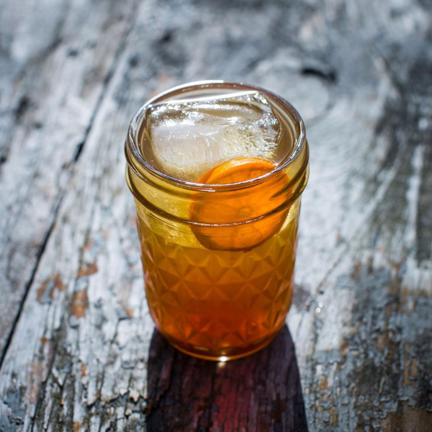 Close-up photo of a light orange cocktail in a small quart-size mason jar with an orange slice and large ice cube in it. The jar is sitting a dark brown rustic wooden table. 